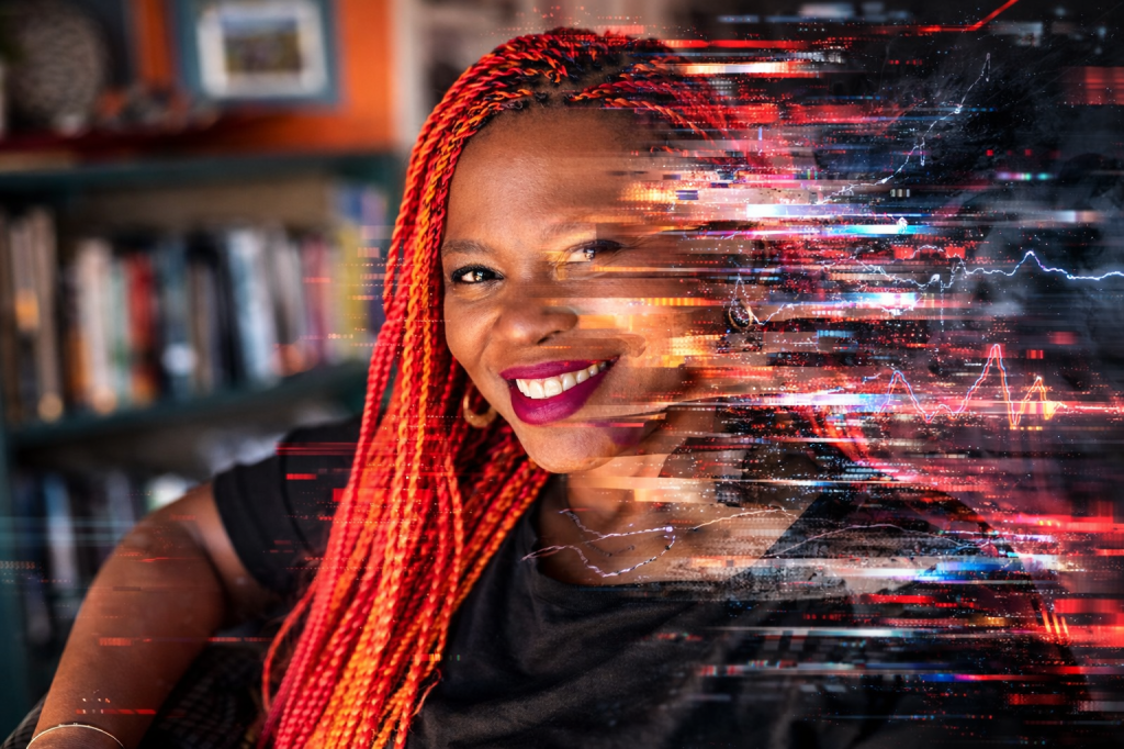 Portrait of a smiling woman with bright red braided hair sitting at a desk, with intense digital glitch distortions across the right side of her face and body, symbolizing invisible neurological disruption.